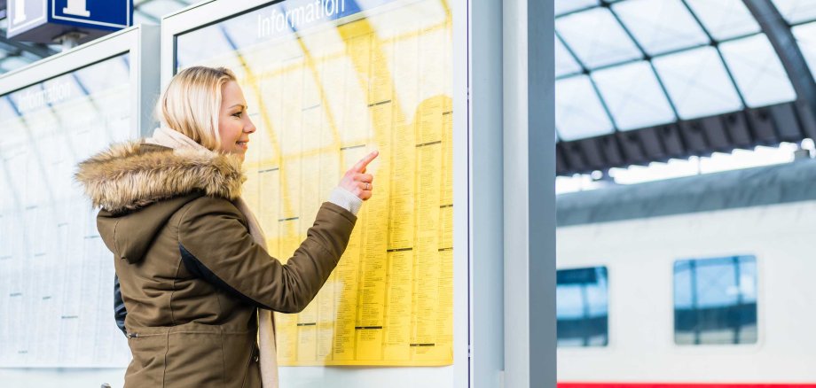Eine blonde Frau in Winterkleidung steht an einem Bahnhof und zeigt auf eine gelbe Informationstafel. Im Hintergrund ist unscharf ein Zug und die Dachkonstruktion der Bahnhofshalle zu erkennen.