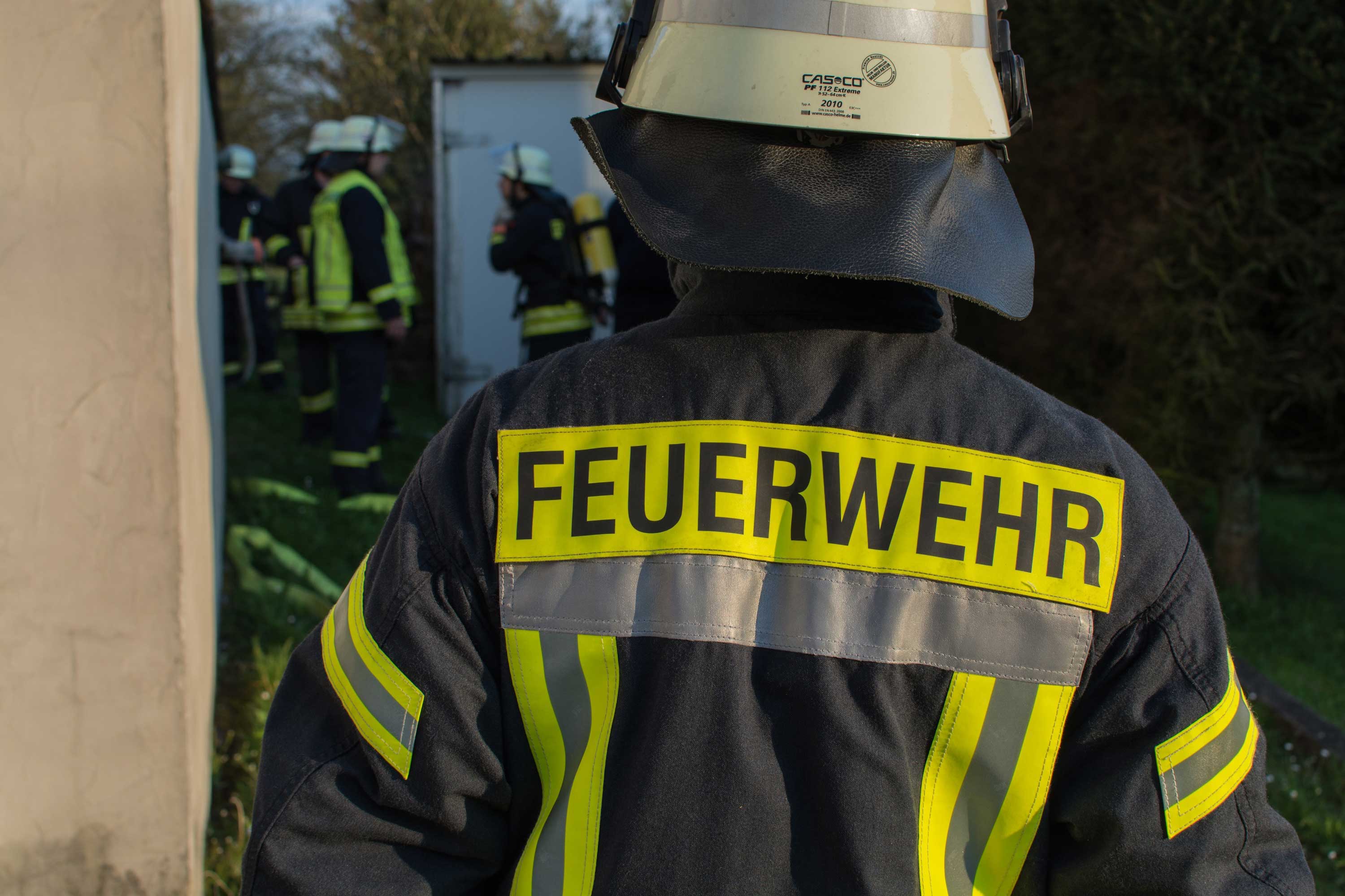 Ein Feuerwehrmann, von hinten gesehen, trägt Schutzhelm und Einsatzjacke mit dem Schriftzug "FEUERWEHR". Im unscharfen Hintergrund sind weitere Einsatzkräfte erkennbar.