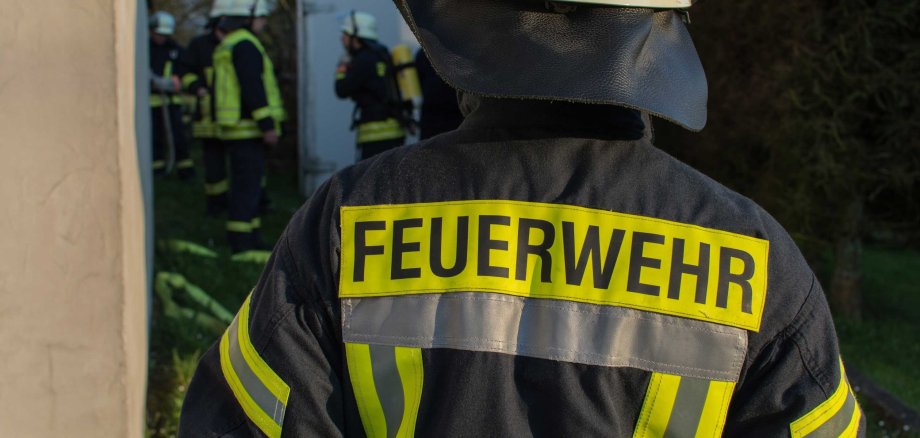 Ein Feuerwehrmann, von hinten gesehen, trägt Schutzhelm und Einsatzjacke mit dem Schriftzug "FEUERWEHR". Im unscharfen Hintergrund sind weitere Einsatzkräfte erkennbar.