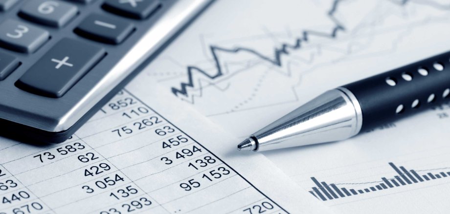 Ein Taschenrechner, ein silberner Stift und Finanzdokumente mit Tabellen und Diagrammen liegen auf einem weißen Schreibtisch. Das Bild symbolisiert Finanzanalyse oder Buchhaltung.