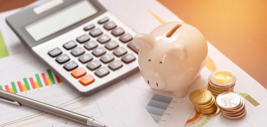 Ein Taschenrechner, ein rosa Sparschwein und Stapel von Münzen liegen neben einem Stift und verschiedenen Diagrammen auf einem Schreibtisch. Sonnenlicht fällt auf die Szene. Symbolisiert Finanzplanung oder Budgetierung.