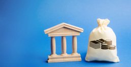 Ein Holzmodell einer Bank mit Säulen steht neben einem Sack mit aufgedruckten Stapeln von Geldscheinen und Münzen. Beide Objekte sind auf einem blauen Hintergrund platziert. Symbolisiert Bankwesen oder Finanztransaktionen.