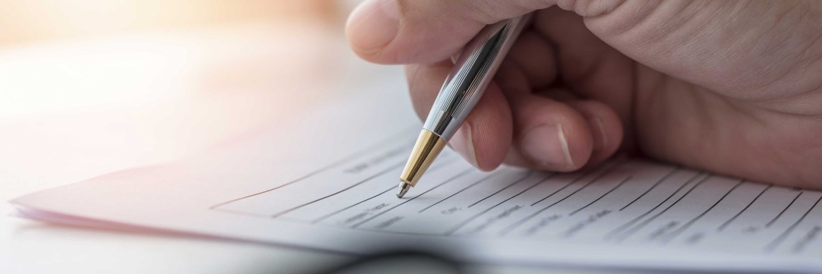 Eine Hand hält einen silber-goldenen Stift und schreibt auf ein Formular mit leeren Zeilen. Sonnenlicht fällt von links oben auf das Dokument und betont die Handlung des Ausfüllens.