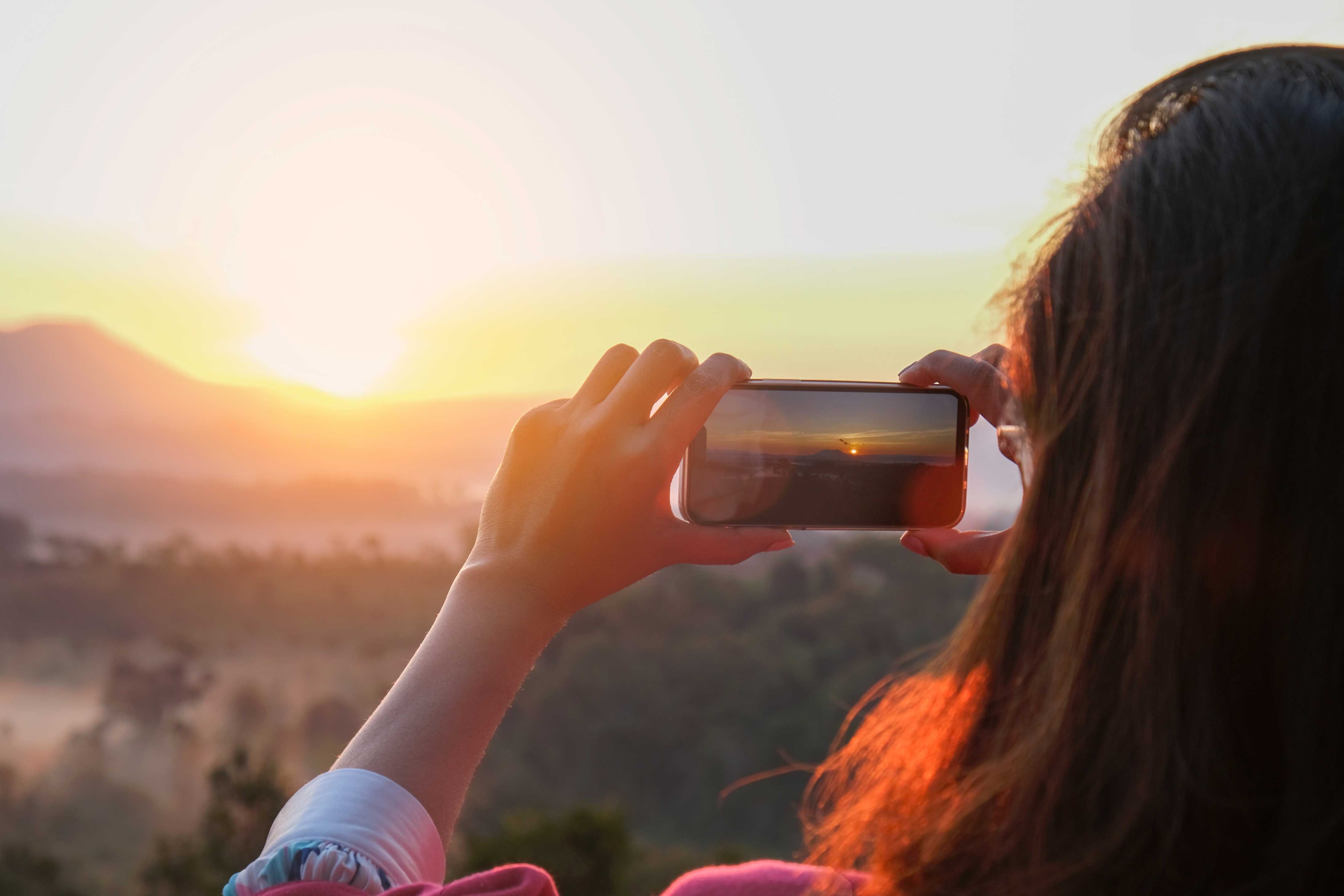 Eine Person hält ein Smartphone und fotografiert oder filmt einen leuchtenden Sonnenaufgang über einer bergigen Landschaft. Das warme Licht spiegelt sich im Haar der Person.