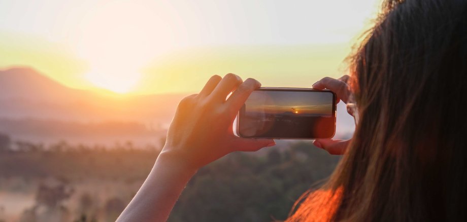 Eine Person hält ein Smartphone und fotografiert oder filmt einen leuchtenden Sonnenaufgang über einer bergigen Landschaft. Das warme Licht spiegelt sich im Haar der Person.