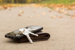 Schlüssel, ein schwarzes Smartphone und eine braune Geldbörse liegen auf einem asphaltierten Weg. Im unscharfen Hintergrund sind herbstliche Blätter auf dem Boden zu sehen.