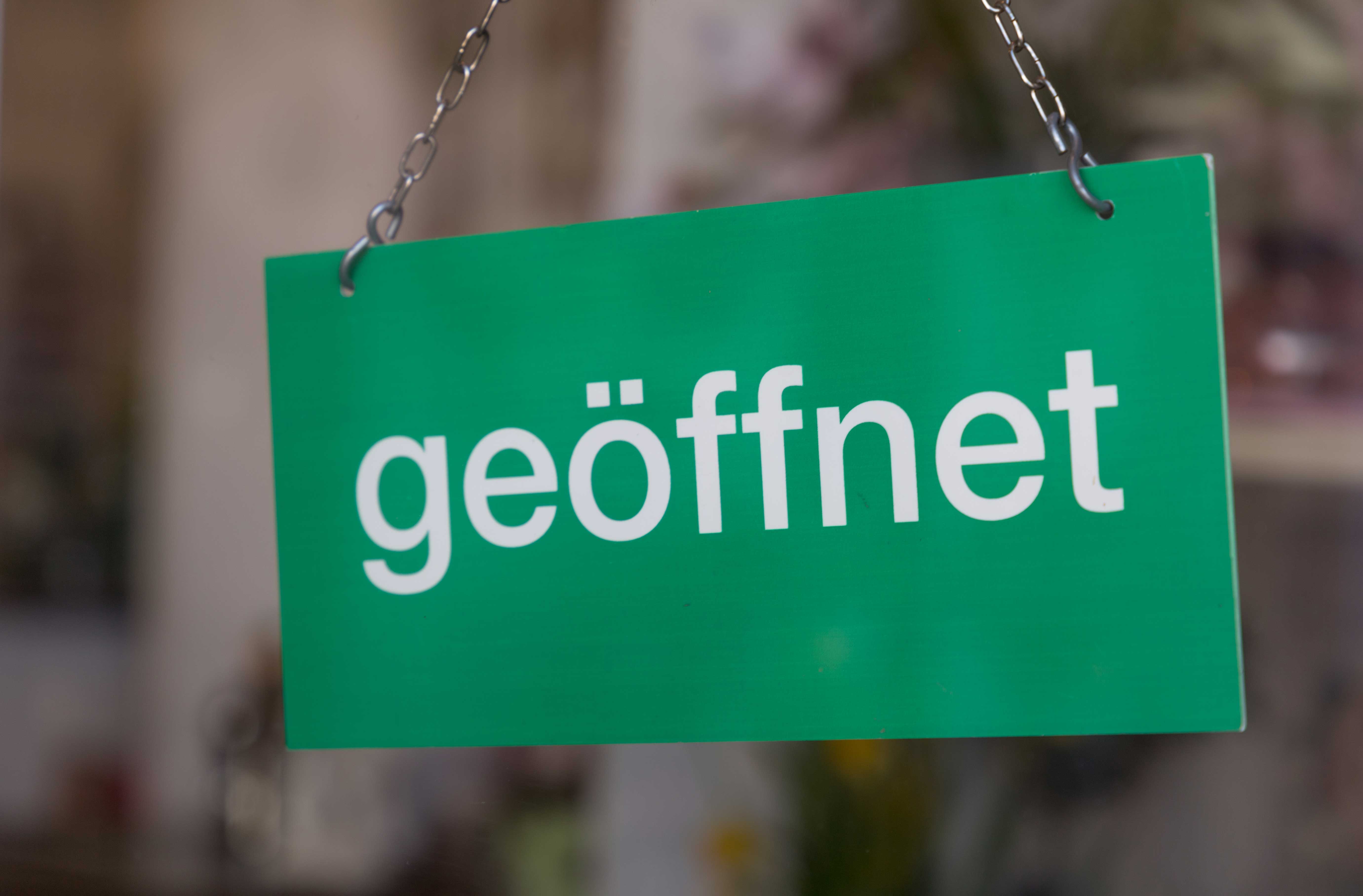 Ein grünes Schild mit der weißen Aufschrift "geöffnet" hängt an zwei Metallketten. Der Hintergrund ist unscharf und deutet auf eine Ladentür oder ein Schaufenster hin.