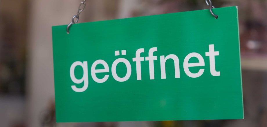 Ein grünes Schild mit der weißen Aufschrift "geöffnet" hängt an zwei Metallketten. Der Hintergrund ist unscharf und deutet auf eine Ladentür oder ein Schaufenster hin.