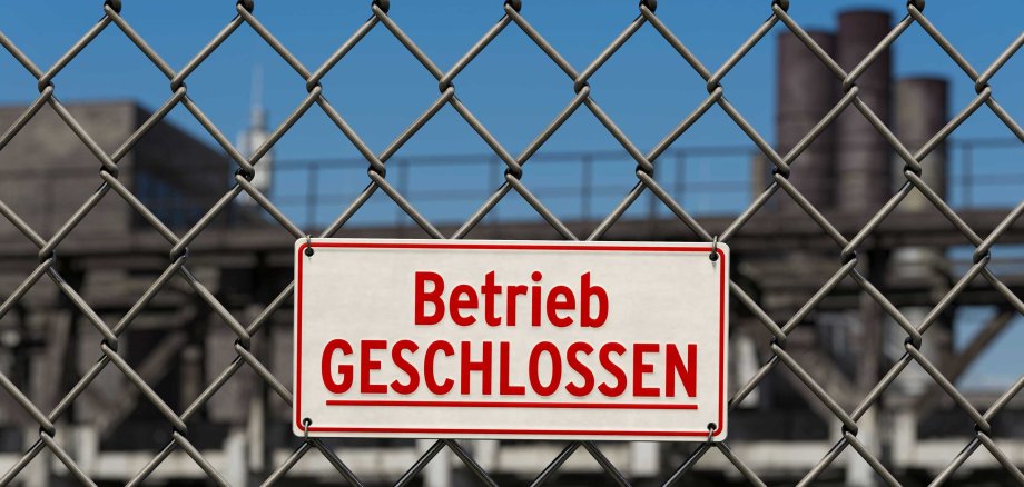 Ein weißes Schild mit roter Aufschrift "Betrieb GESCHLOSSEN" ist an einem Maschendrahtzaun befestigt. Im unscharfen Hintergrund sind Fabrikanlagen zu erkennen. Symbolisiert eine Stilllegung oder Insolvenz.